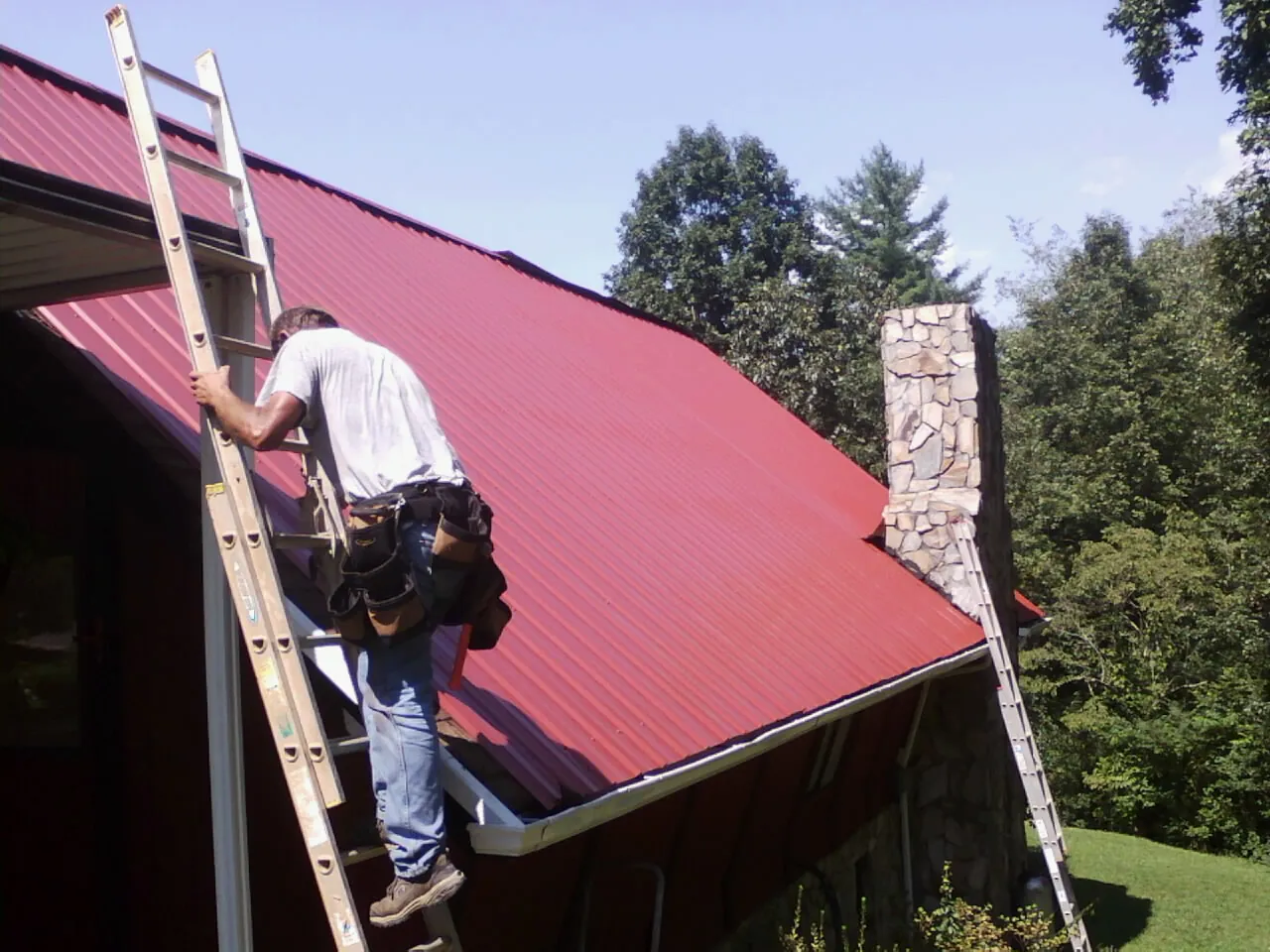 Roofer on a ladder beside a red metal roof and stone chimney — Soffit & Fascia Repair by Roofing Brothers in Whitefish